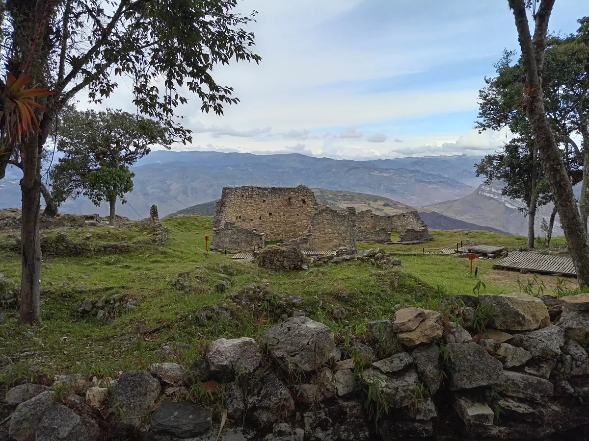 The partially standing curved stone walls of a circular house ruin at Kuélap, rising to around head height on a grassy hilltop with the Amazonas mountains behind